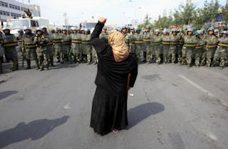 Woman on crutch shouts at Chinese paramilitary police wearing riot gear as crowd of angry locals confront security forces on street in Urumqi